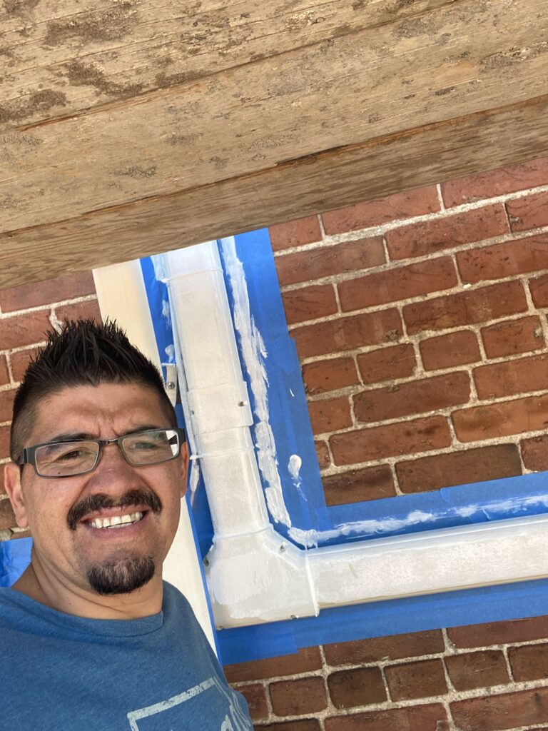 Owner Alex Muniz on a job site beside a window frame being sealed and painted on a brick building
